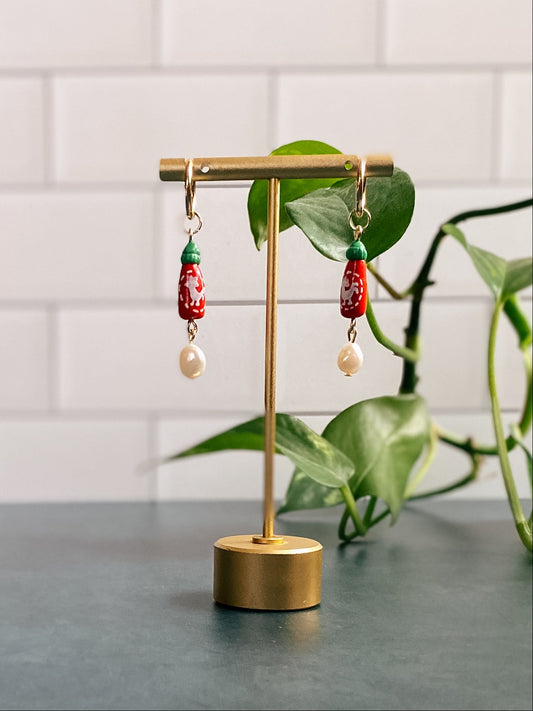 Gold earring stand with red and green sriracha bottle earrings with a pearl on a white tiled wall background